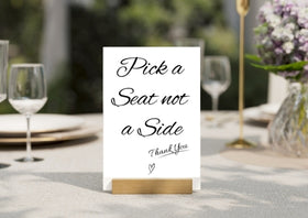 Pick a Seat not a Side A5 wedding sign displayed in a gold metal stand on a white table with floral background