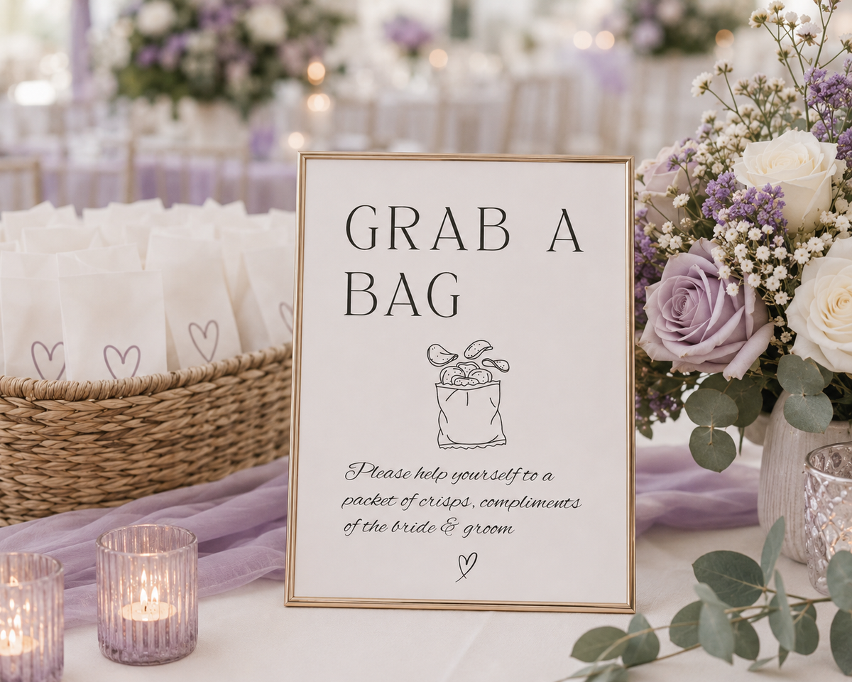 A wedding table sign reading 'GRAB A BAG' with a small gift box illustration and handwritten text below, styled with a woven basket, white napkins, lavender candles, and floral arrangements in soft purple, white, and green tones.