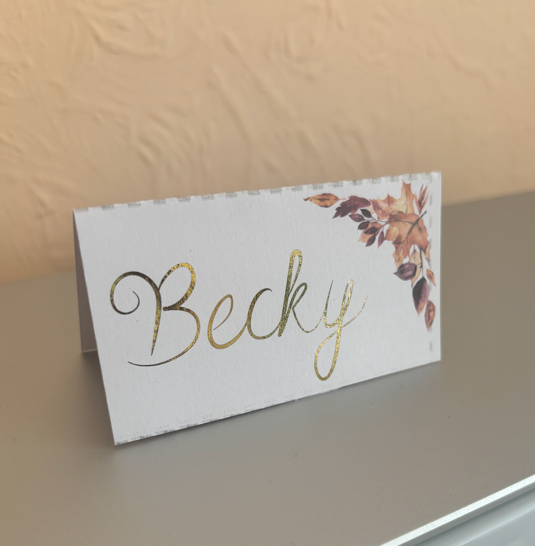 A personalized place card with the name 'Becky' in gold foil script and a watercolor autumn leaf design, standing on a gray surface against a beige wall.
