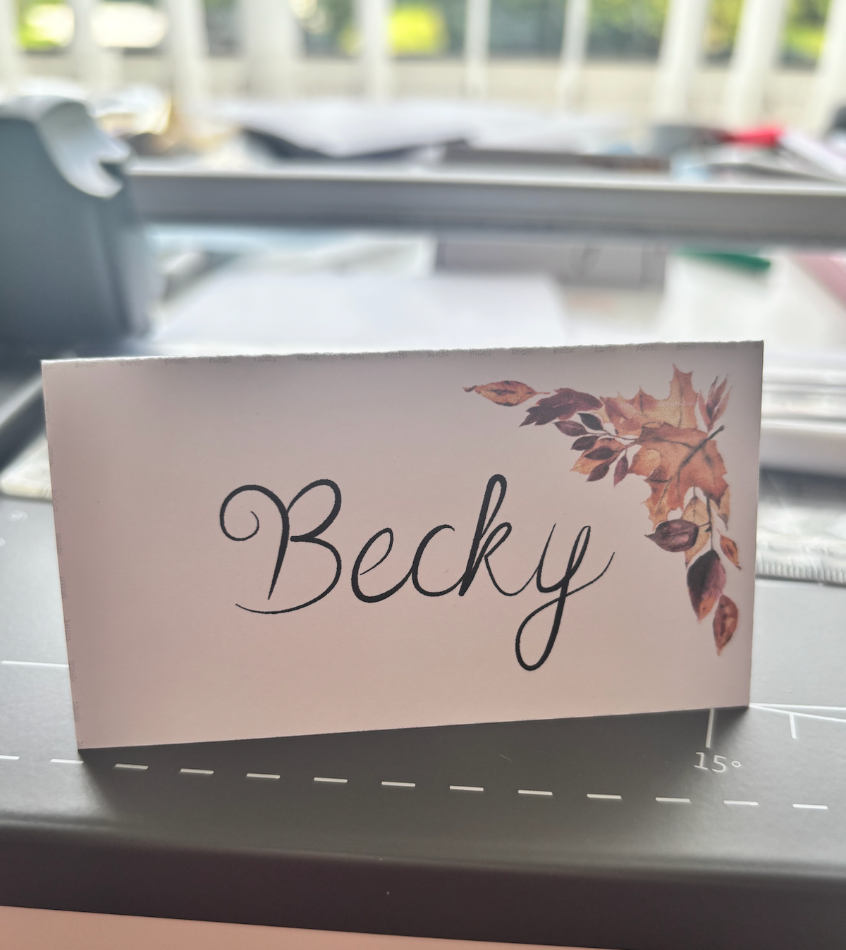 A rectangular white table name card with the name 'Becky' in black script and a cluster of autumn leaves in brown and orange at the top right, photographed on a dark tabletop with a blurred indoor background.